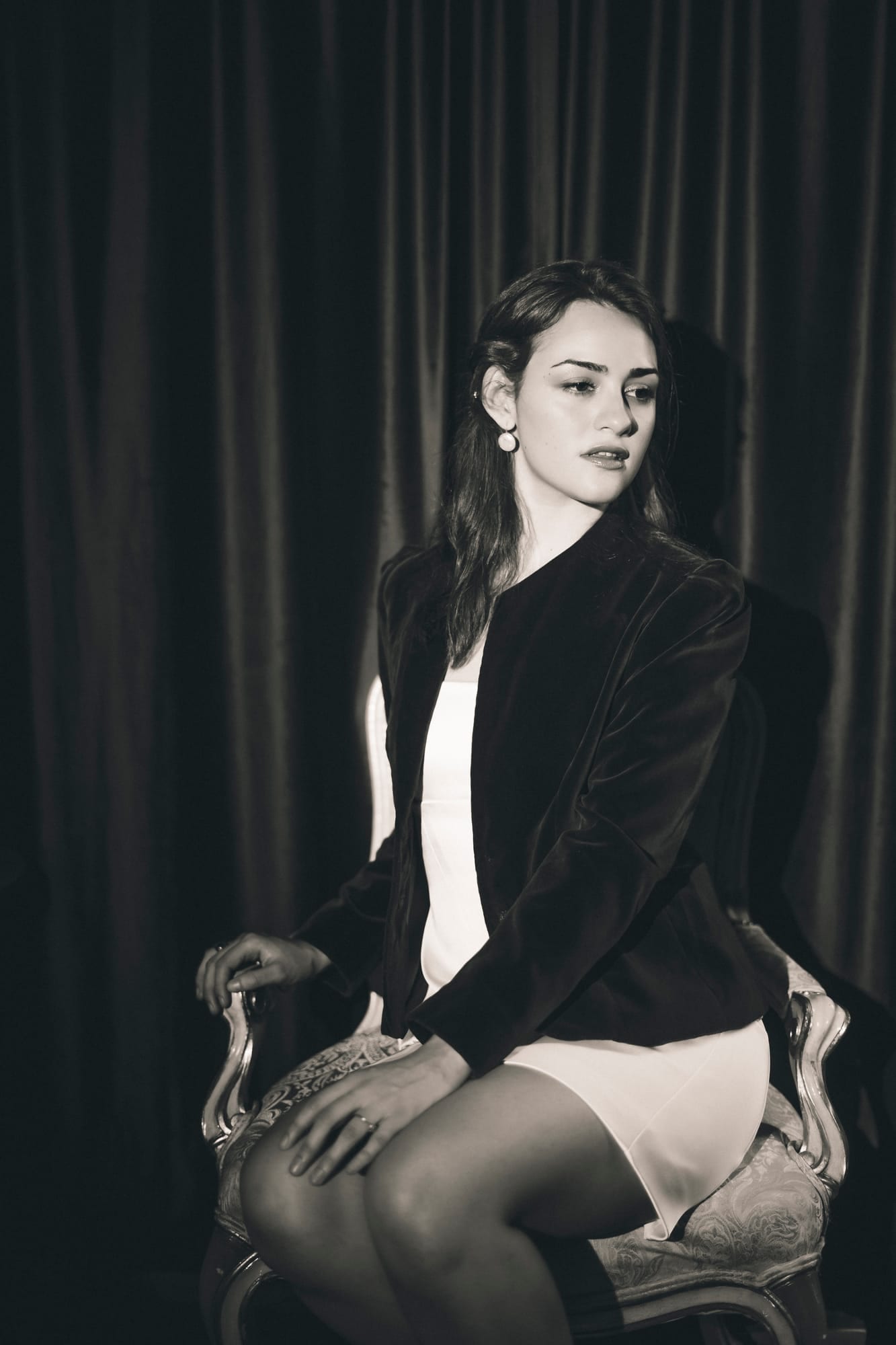 Seated in a velvet jacket against a dark curtain